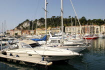 Boats docked with tall masts. Buildings in front of wooded area in the distance.