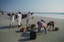 Three men in turbans watch three women sorting the morning catch near a boat on the beach.