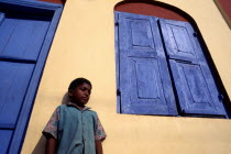 Angled view of a boy standing by a yellow house with a blue door and window shutters