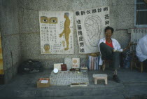 Roadside medicine seller seated in front of medical drawings of the human body