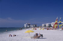 Woman reading under a multicoloured parasol on a sandy beach with hotels in the distance