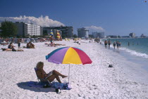 Sunbathers on a golden sandy beach with a yellow parasol and sailboats along the beach behind