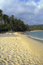 View along golden sandy beach lined with palms and bathers by boats pulled on to the sand