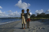 Two children standing on shoreline of sandy beach.