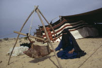 Bedouin woman with baby in suspended cot made from animal hide with tent behind.