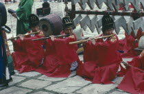 Confucian Rites Orchestra. Line of men in red robes sat on ground playing bamboo flutes