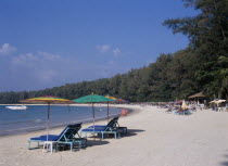 View along the sandy beach with sun loungers and colourful parasols in the foreground and a row of them all along the water s edge.. Red sign for boat hire and people swimming  boat in the water. Col...