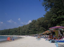 View along the sandy beach with row of sun loungers and colourful parasols. Red sign for boat hire and children playing and digging in the sand. Colorful