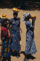 Girls wearing brightly coloured dresses selling tomatoes carried in bowls on their heads. Colored