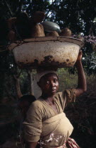 Portrait of young woman carrying bowl of yams on her head and baby in sling on her back.