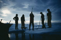 Silhouette of a man using an axe to cut through ice. Others stand round  two holding spears.