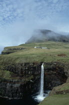 Waterfall and rocky coastline with houses built on green landscape