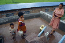 Woman drawing water from pump on roof of building.