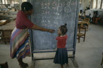 Female teacher pointing out 2 x table written in coloured chalk on blackboard to little girl.