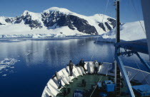 View of the front section of a ship with people standing looking over edge surrounded by snow covered mountains