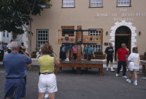 Tourists in kings Square with people in the foreground talking pictures of a man standing in a wooden execution guillotine outside The Bank of Butterfield Building