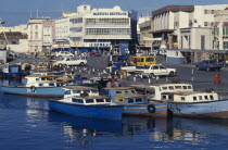 Moored painted boats on the Careenage with busy shopping district beyond.