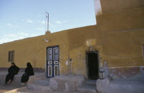 Two Muslim women seated outside yellow coloured building.