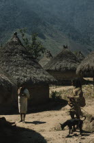 San Miguel village  important Kogi centre  thatched roofs with pots at apex.  Man with dog wearing threadbare woven  manta  / cloak and two  mochilas  / shoulder bags  talking to woman with new  manta...