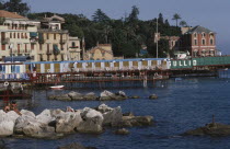 Coastal town on the Italian Riviera with line of changing huts on jetty out into rocky bay wand waterside buildings behind.