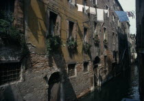 Canalside houses with crumbling plaster and brickwork  plants and flowers in window boxes and washing hanging between buildings overhead.