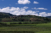 Road to Ambalavao. View across green fields with people leading Zebu cattle along a pathway dividing paddy fields with thatched huts in hillside village