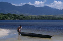 A woman standing at the waters edge of a sandy beach about to push her Pirogue back into the water to return to her village with mountains in the distance