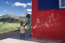 Road to Fianarantsoa. Toddler with Grandmother appearing from around exterior wall of a roadside cafe with Classiko red and blue livery and lush green landscape and hills in the distance behind them