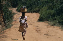 Near Ambositra. Portrait of a woman carrying a baby in a sling on her back and wicker baskets on her head walking up a red earth road