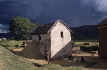 Road to Ambositra. Women outside thatched white building with straw in a pile next to the wall of the building and dramatic dark cloud formation in the sky