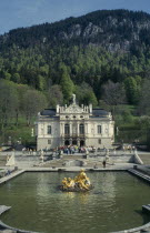 Palace exterior with tourists crowds gathered at entrance.  Ornamental lake with gold statue in the foreground.  Smallest of three castles built by King Ludwig II. Bavaria