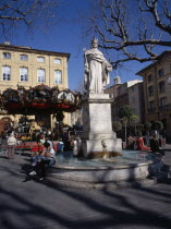 Statue in centre of water fountain  people sat on edge. Carousel and buildings behind. Center