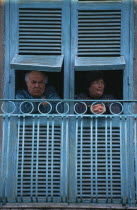 San Giorgio a Cremano.  Elderly couple looking out from turquoise open wooden window shutters above street.