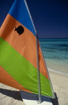 Sail board with colourful  striped sail pulled up on sandy beach with sea behind.