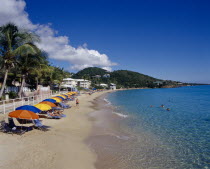 Frenchman Bay.  View along sandy beach with row of sun loungers and brightly coloured beach umbrellas. People sunbathing and swimming.