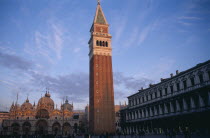 The Piazza San Marco with the Campanile bell tower in the foreground and the Basilica di San marco behind.  Crowds of people and pigeons.