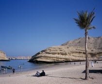 Bandar Jissah beach near Al-Bustan Hotel just outside the capital city.  People at water s edge beside eroded  rock formation with palm in foreground.