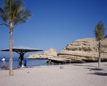 Bandar Jissah beach near Al-Bustan Hotel just outside the capital city.  People at water s edge beside eroded  rock formation with sun umbrella and palm trees.