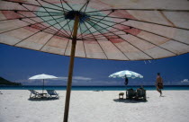 Western tourists on white sand beach with sun loungers part framed by umbrella in the foreground.