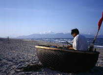 Fisherman mending nets in coracle boat pulled up onto sandy beach.