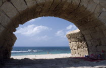 Roman Aqueduct with view through a single arch towards sunbathers on the beach next to the Mediterranean Sea