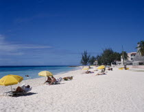 Golden sandy beach lined with palms  with scattered bathers  sunbeds and umberellas and buildings partially hidden by trees.