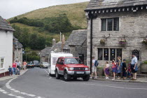 People walking along pavements next to traditional cottages by a road with passing traffic and caravansGreat Britain United Kingdom UK