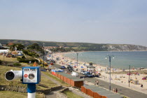 Elevated view across beach huts towards busy sandy bay with sunbathers. Viewing telescope in the foreground.Great Britain United Kingdom UK