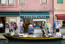 A fruit and vegetable vendor with his boat moored alongside the Fondamenta dei Vetrai olong the Rio dei Vetrai canal on the lagoon island of Murano. Tourists walk along the pavement looking at the sho...