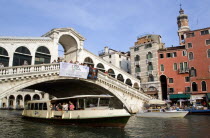 A vapretto passes below the Rialto Bridge with tourists viewing the canal