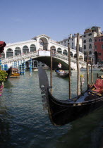 A gondola moored at the edge of the Grand Canal with another carrying tourists passes down river in front of The Rialto Bridge with tourist crowds