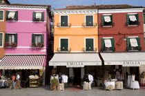 Brightly coloured houses above lace shops on the lagoon island of Burano the historic home of the lace making industry in the region. Tourists walk past looking at the displays on the pavement Colore...