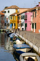 Colourful houses beside a canal on the lagoon island of Burano with people walking past boats moored alongside the edge of the canal Colorful