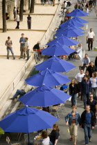 The Paris Plage urban beach. People strolling between the River Seine and people playing boules or petanque divided by a line of tables under umbrellas along the Voie Georges Pompidou usually a busy r...